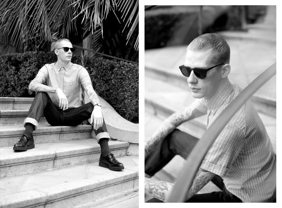 A man sits on stairs in sunglasses wearing a 3sixteen short sleeve shirt.
