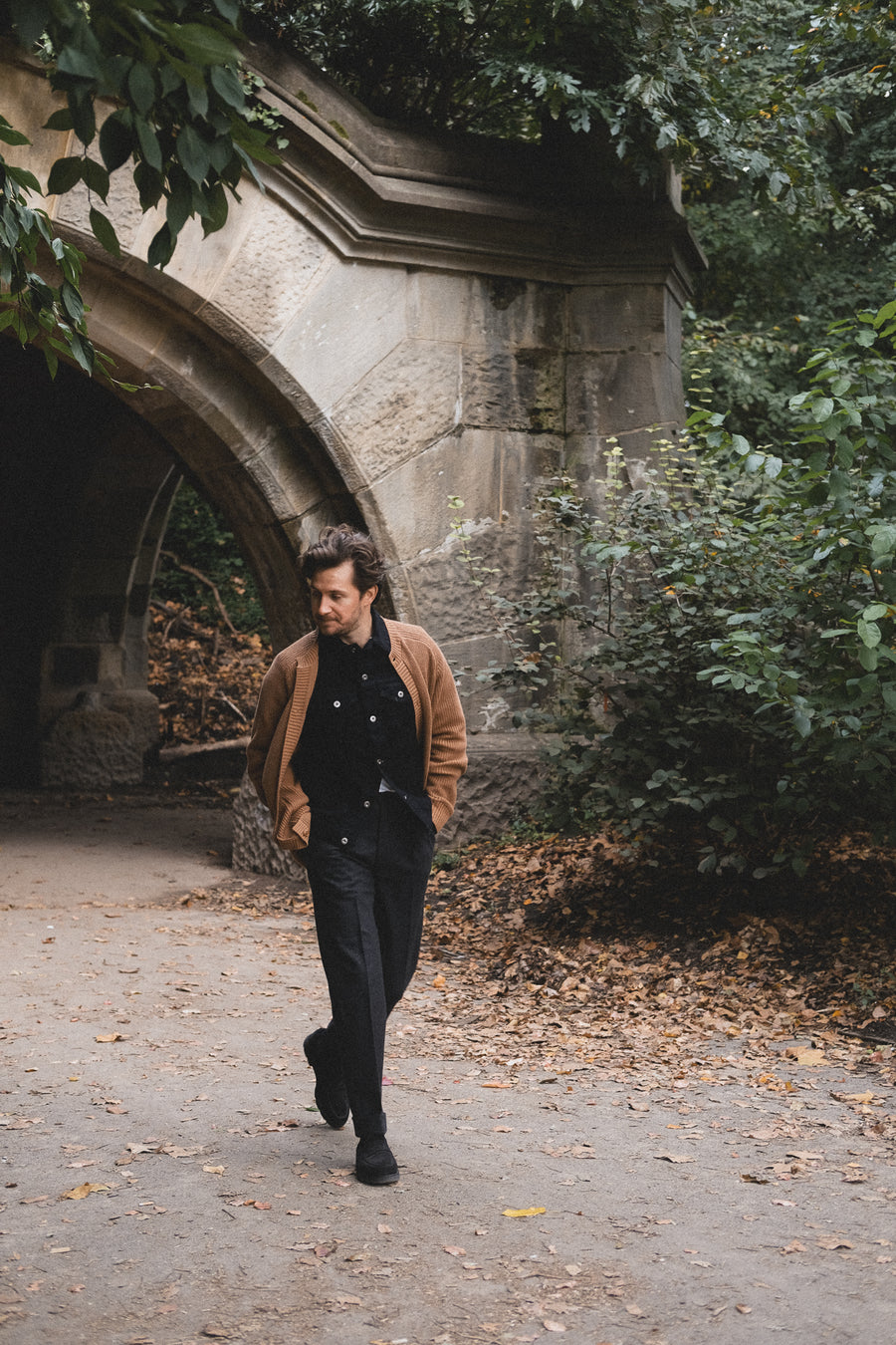 A man walks out of a stone archway in a brown sweater.