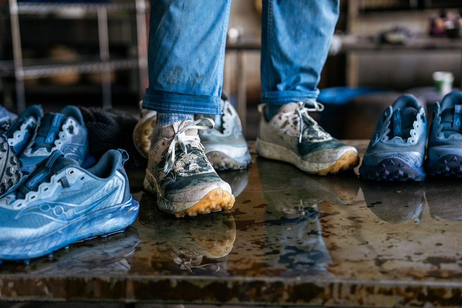 A person wearing a pair of indigo-stained white shoes stands amongst pairs of indigo dyed shoes on a metal table.