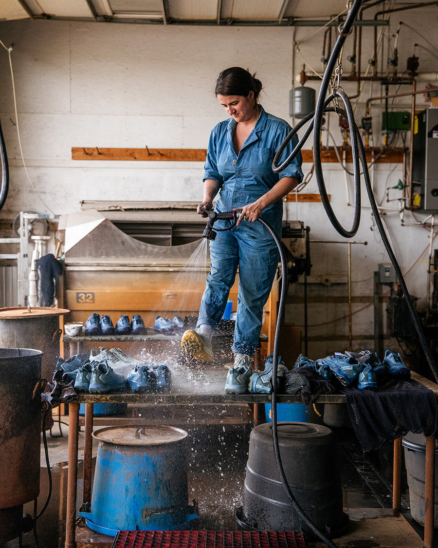 A woman in a blue jumpsuit hoses down sneakers on a table.
