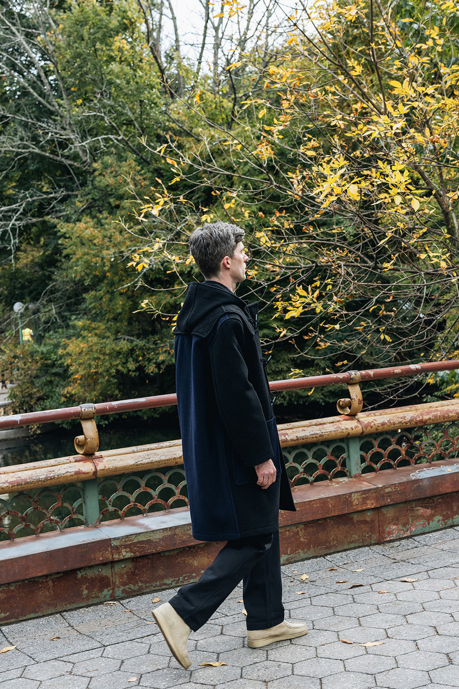 A man in a black wool coat walks across a bridge in a park.
