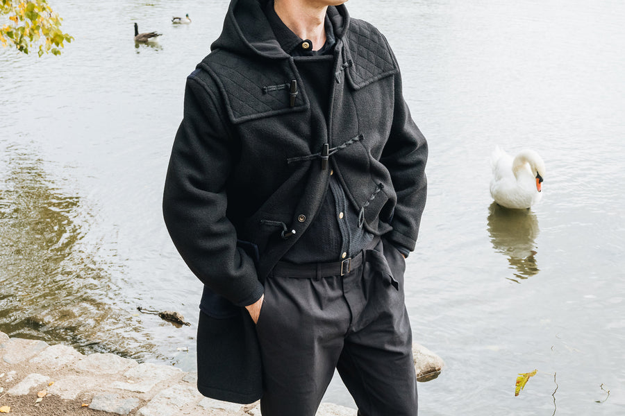 A detailed shot of a black wool overcoat worn next to a lake with a swan in it.