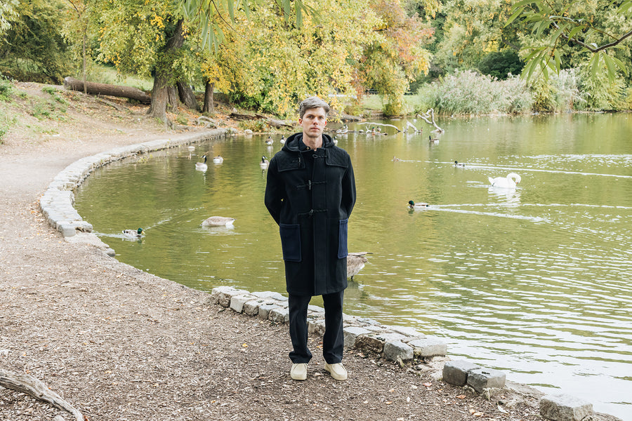 A man in a black wool coat stands by a lake.