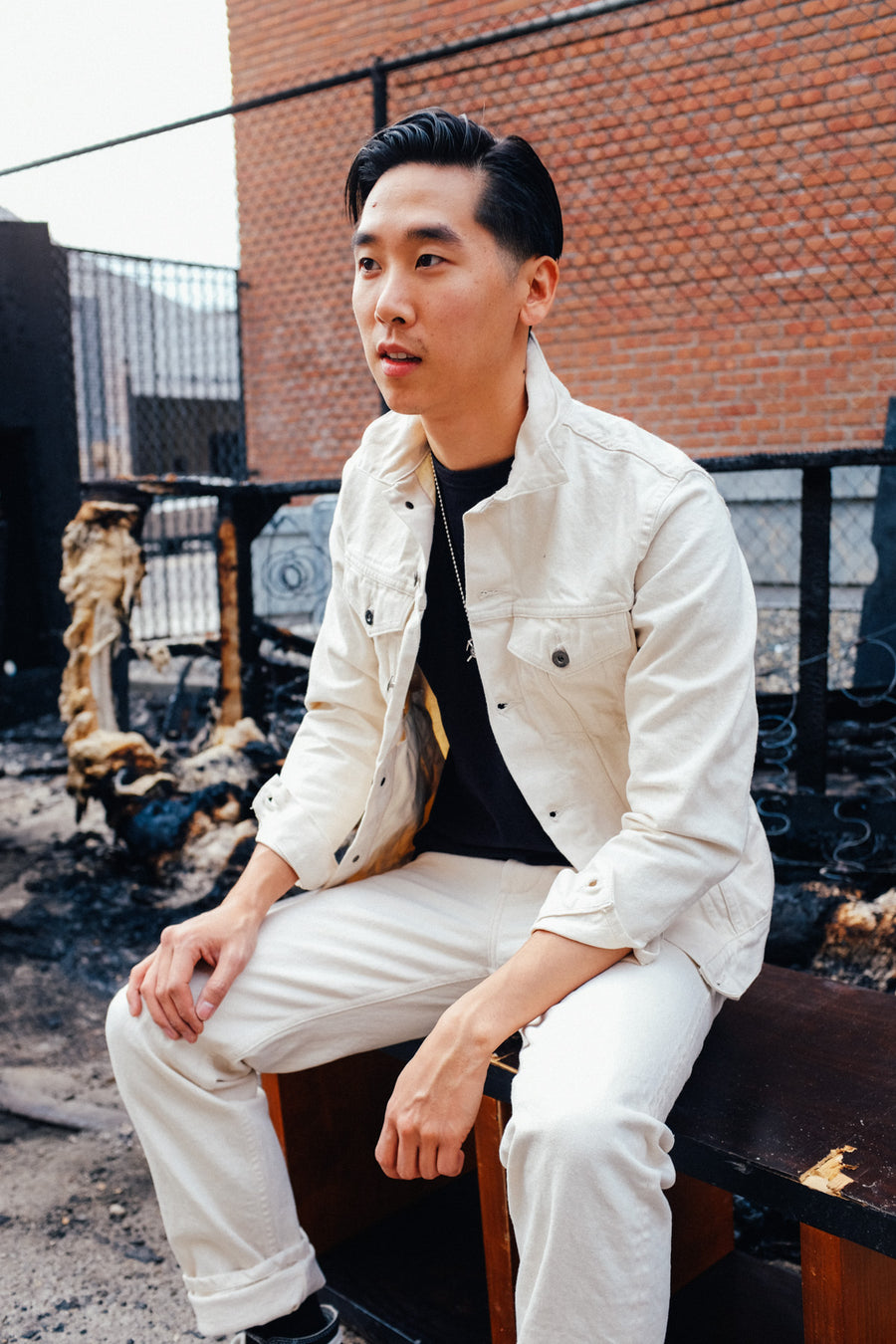 A young man sits on a broken bookshelf wearing an ecru denim jacket and chinos.