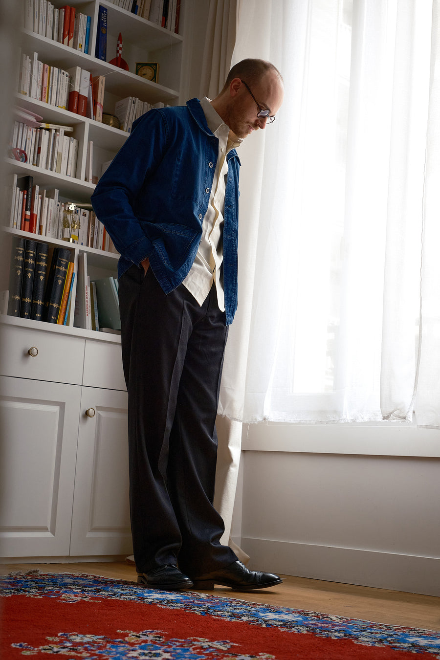 A man in a blue jacket, white oxford and wool pleated pants stands in a living room.