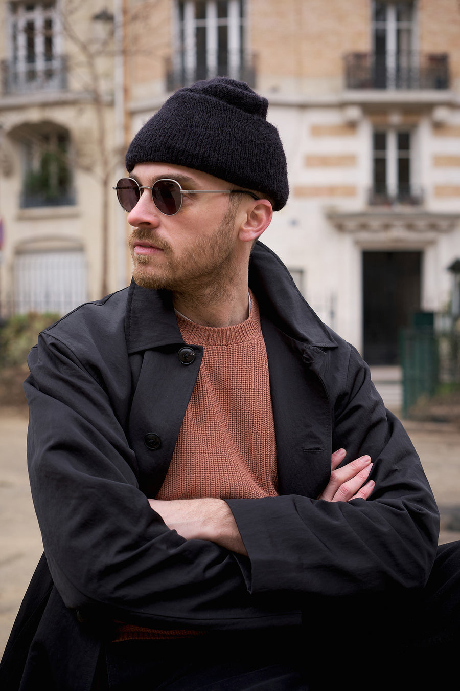 A man in a black hat and coat with an orange sweater sits on a park bench.