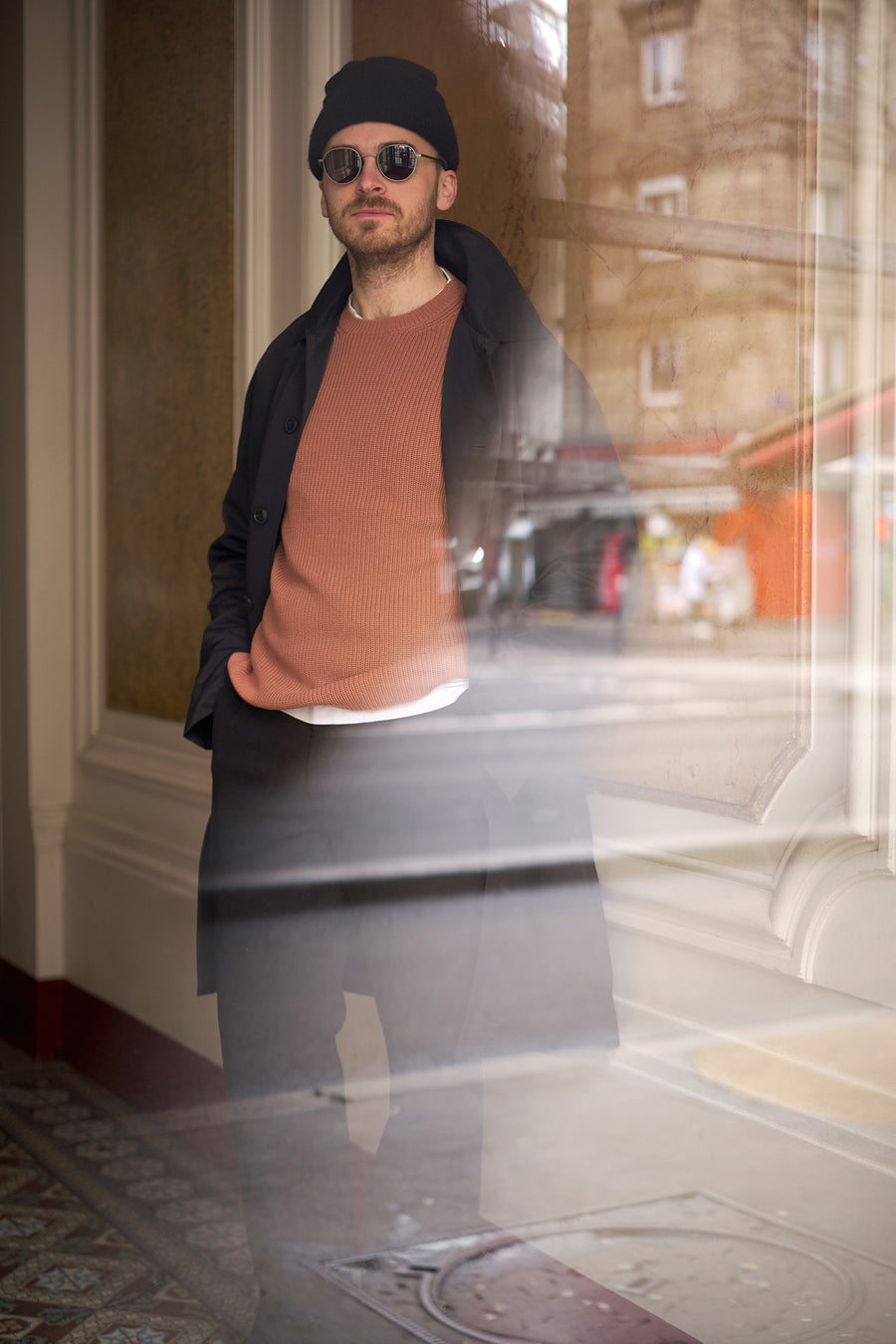 A man in sunglasses and a black coat stands behind a glass door.