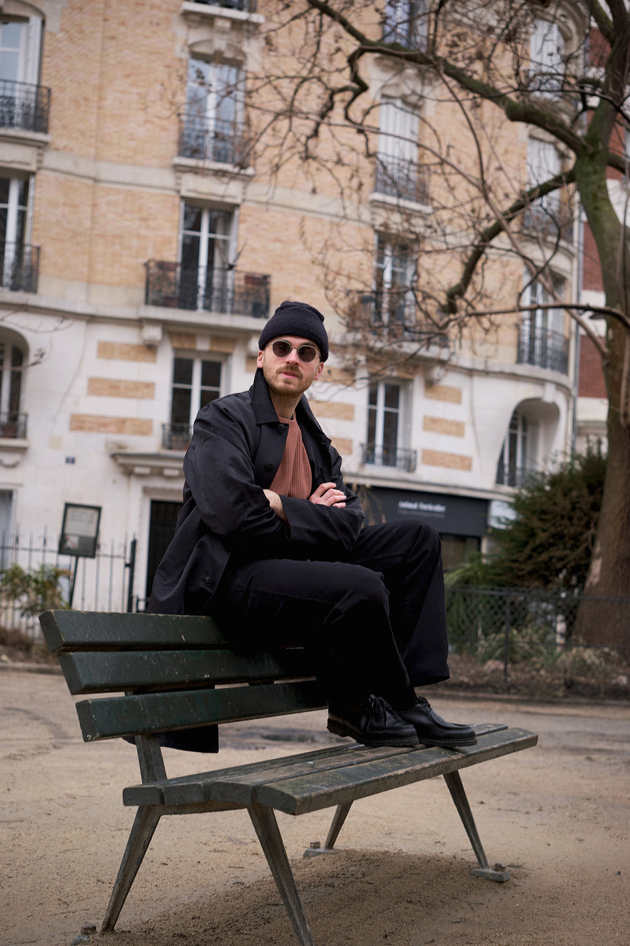 A man in a black coat and orange sweater sits on a park bench.