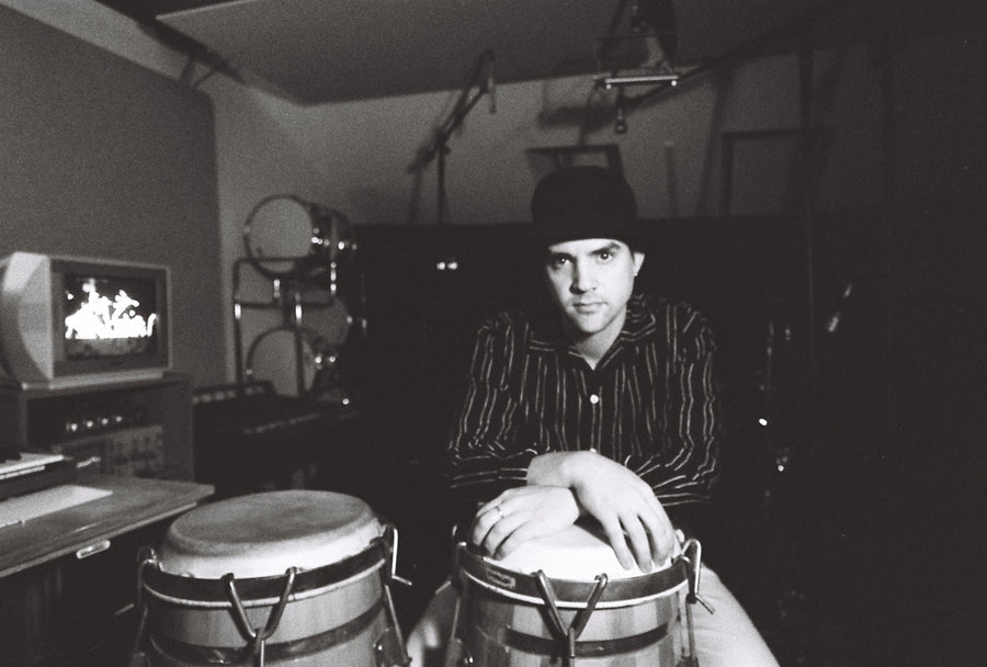 A man in a striped shirt sits with some congas.