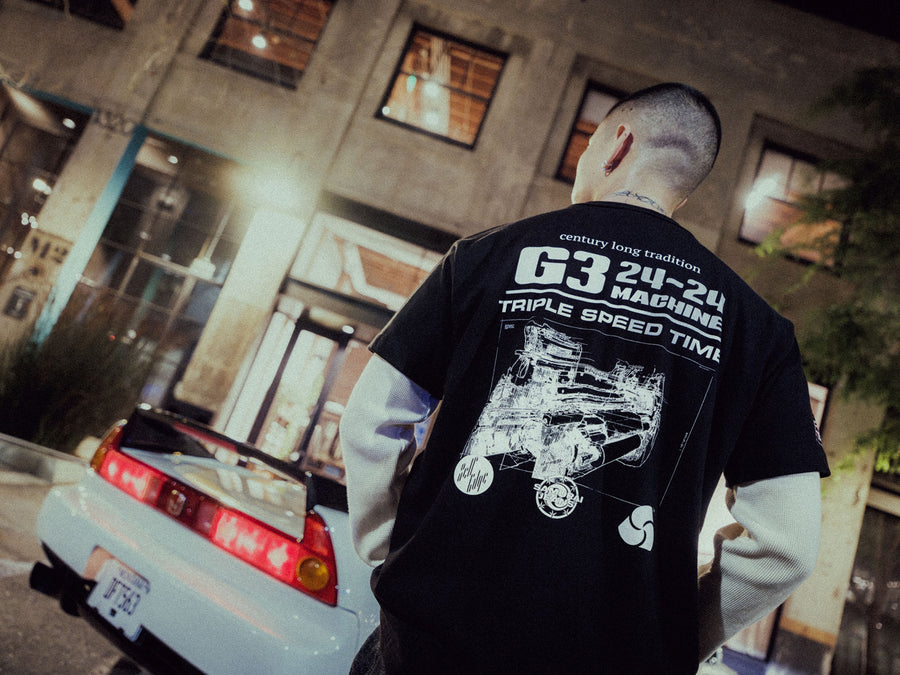 A man with a graphic tee with a selvedge denim loom on the back of it stands in front of a car.