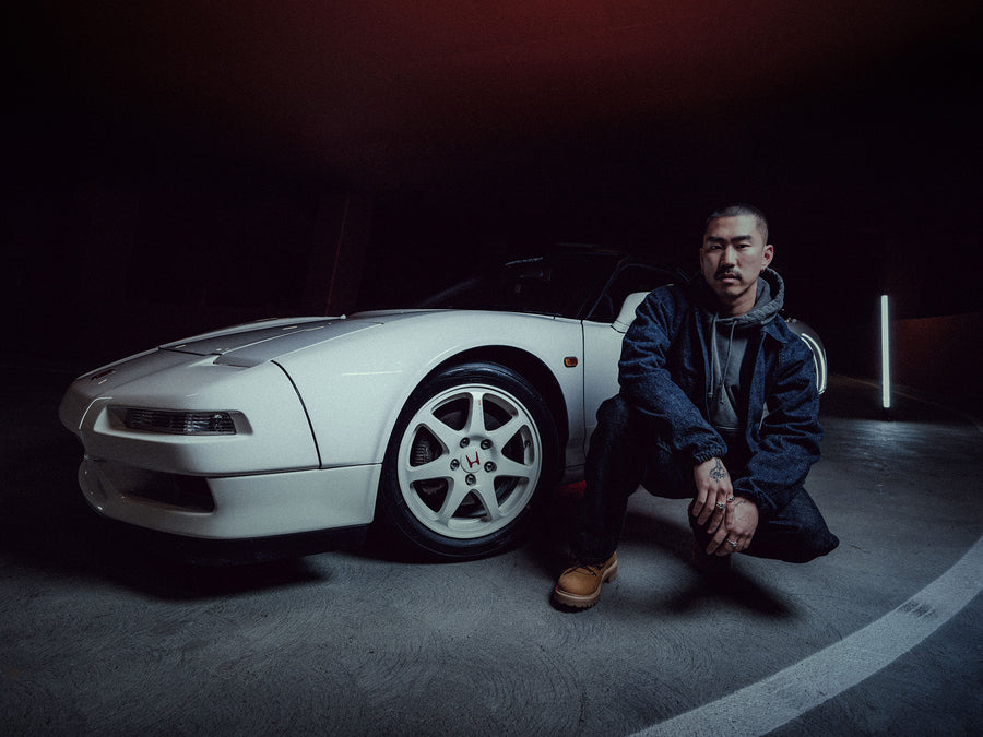 A man in a denim jacket and jeans poses by a vintage Honda sports car.