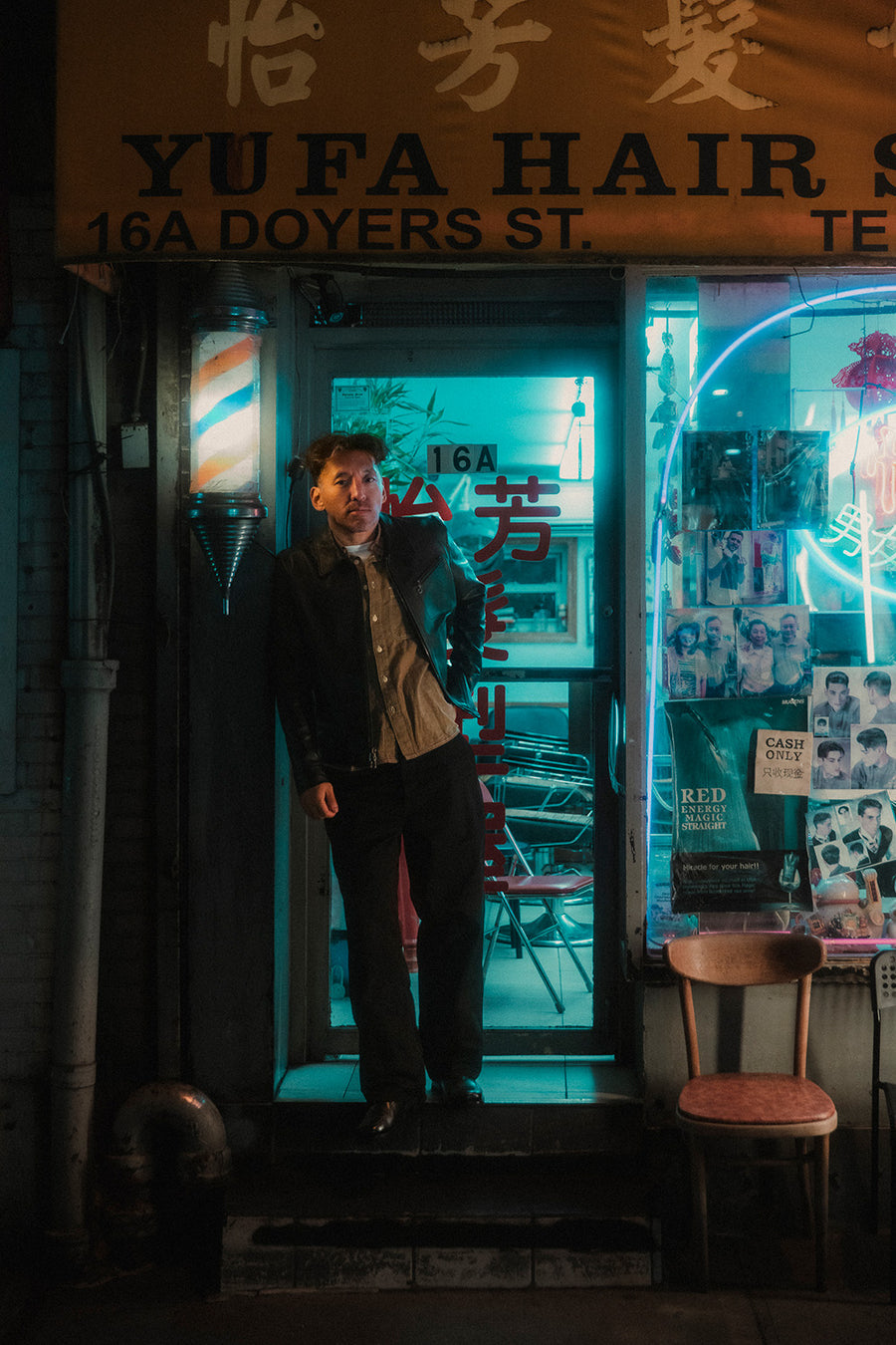 A man stands in front of a barbershop in a black leather jacket.