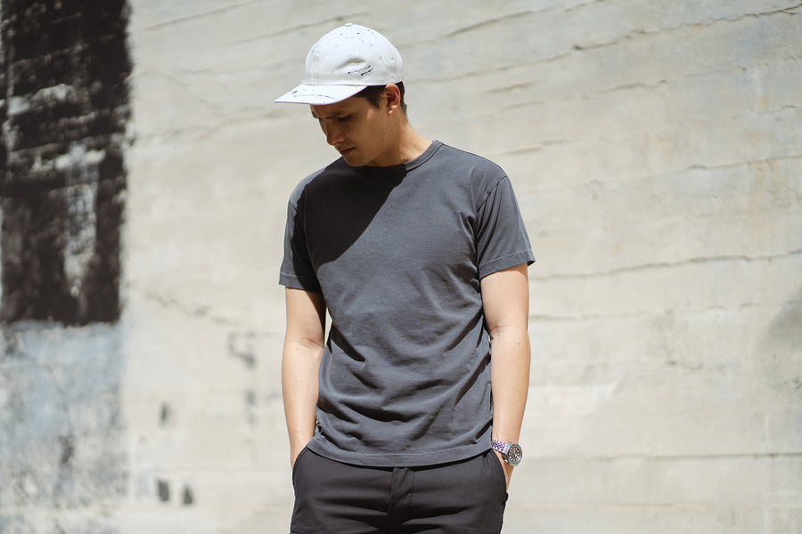 A man in a white baseball cap and the smoke garment dyed tee stands in front of a white wall.
