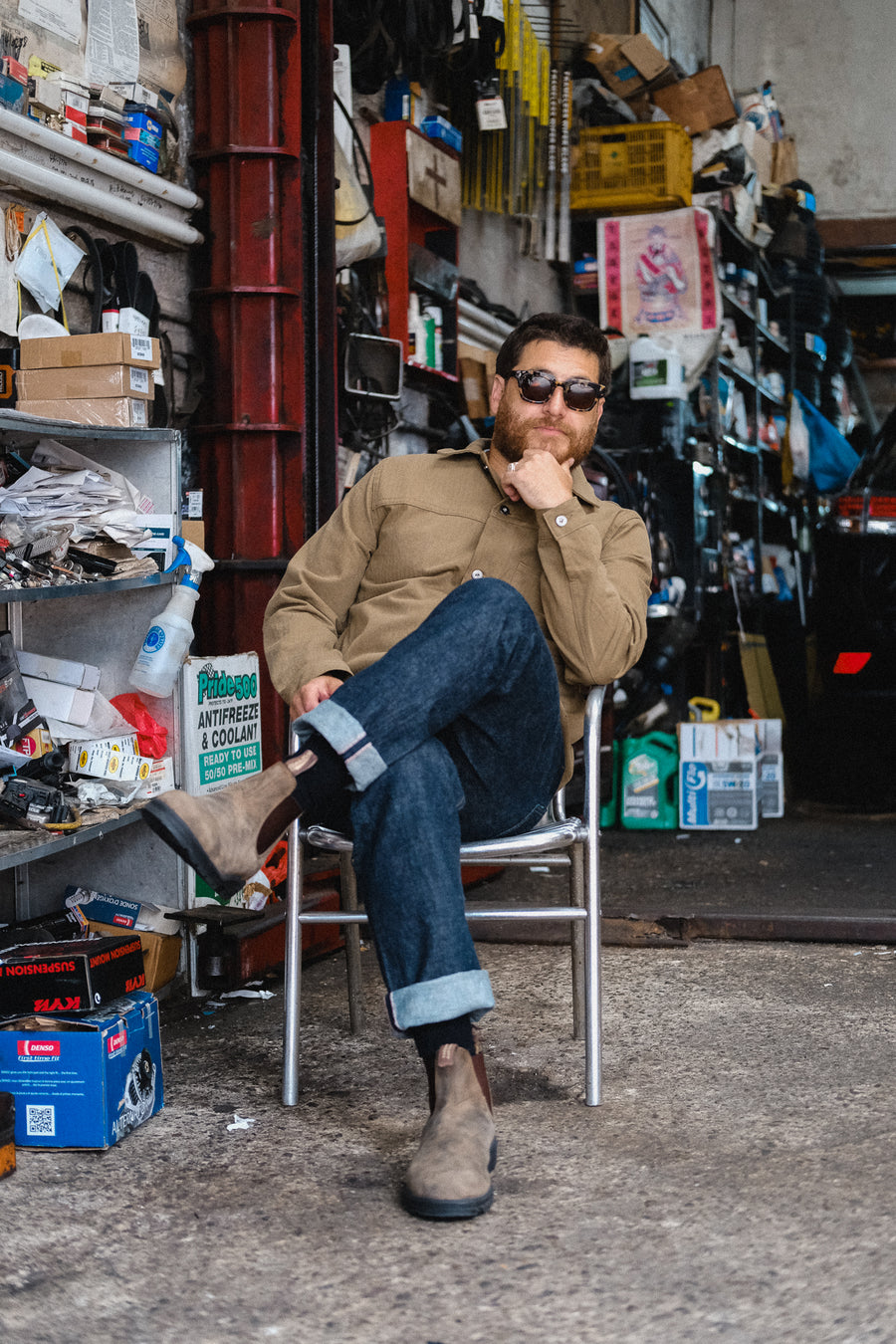 A man in a tan jacket and jeans sits on a chair in a garage.