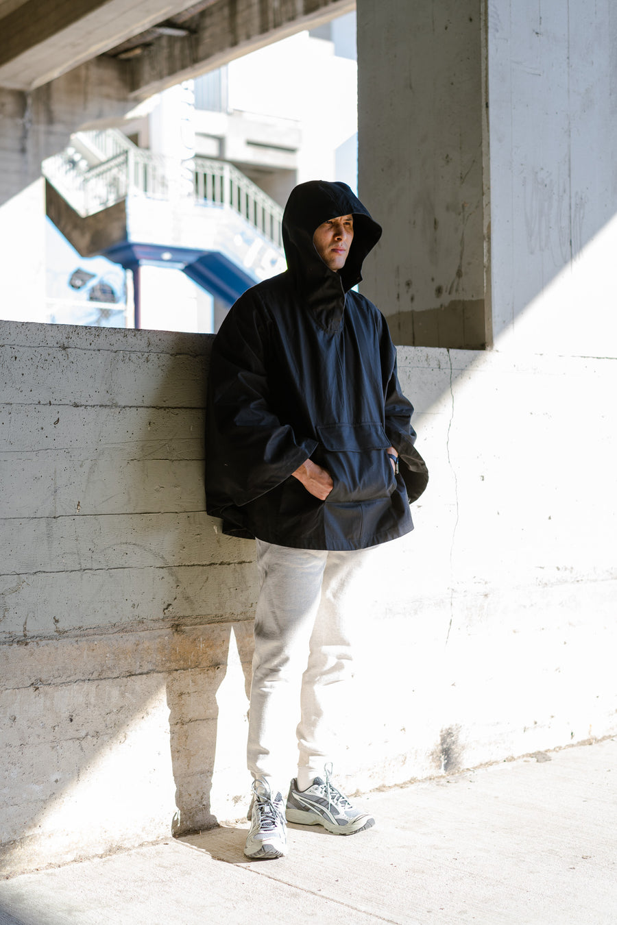 A man in a black poncho and sweatpants stands under an overpass.