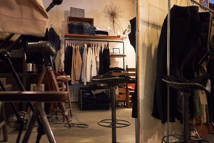 A photo of a store as seen through a mirrors reflection. Warm, yellow tones and pipe racks hold clothing.