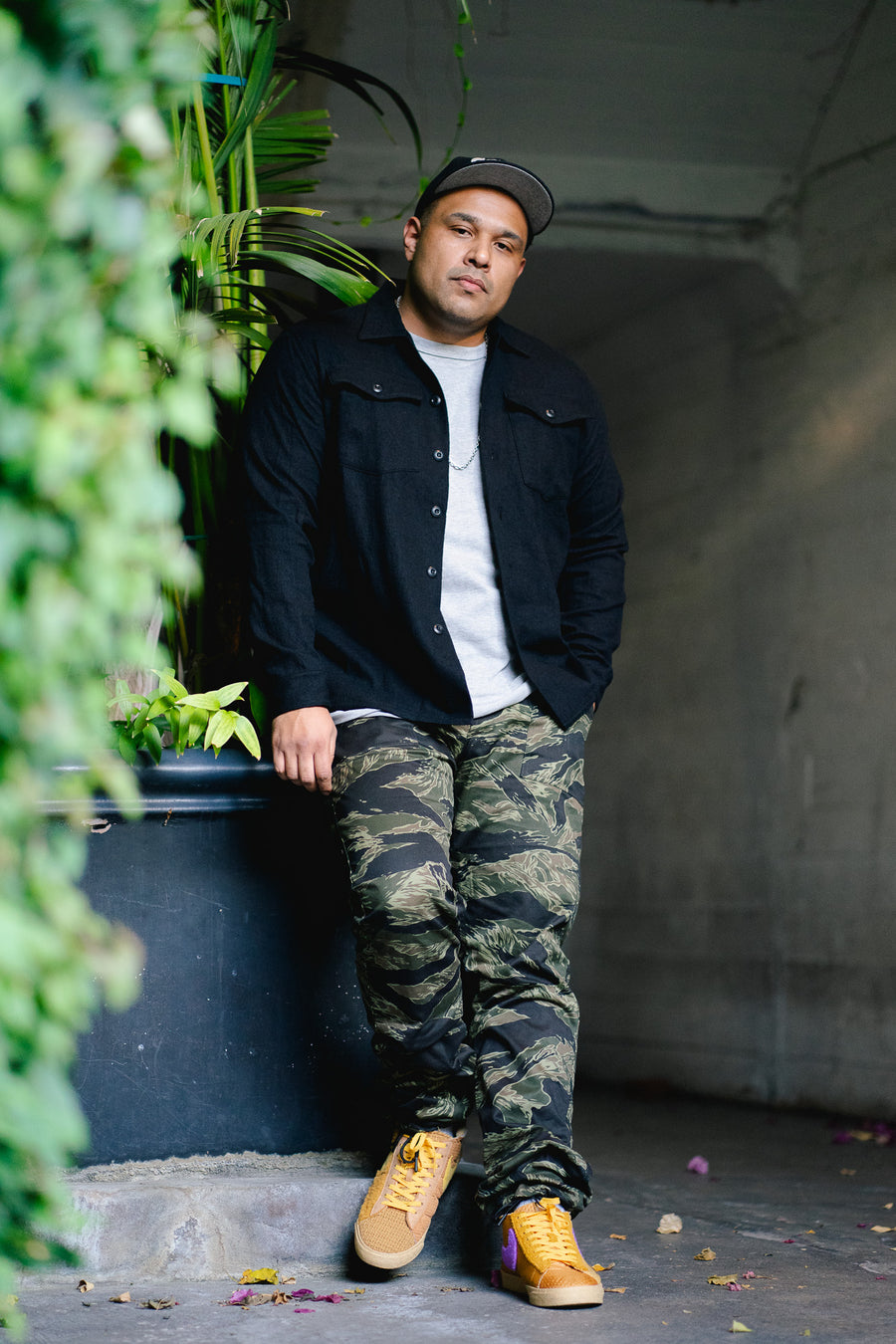 Man leans against a large planter in a black shirt and camouflage pants