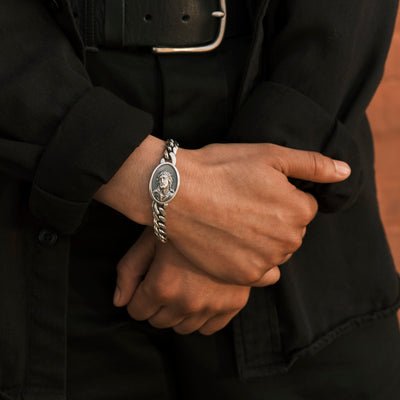 A silver bracelet with a Jesus head medalliaon is worn on a wrist.