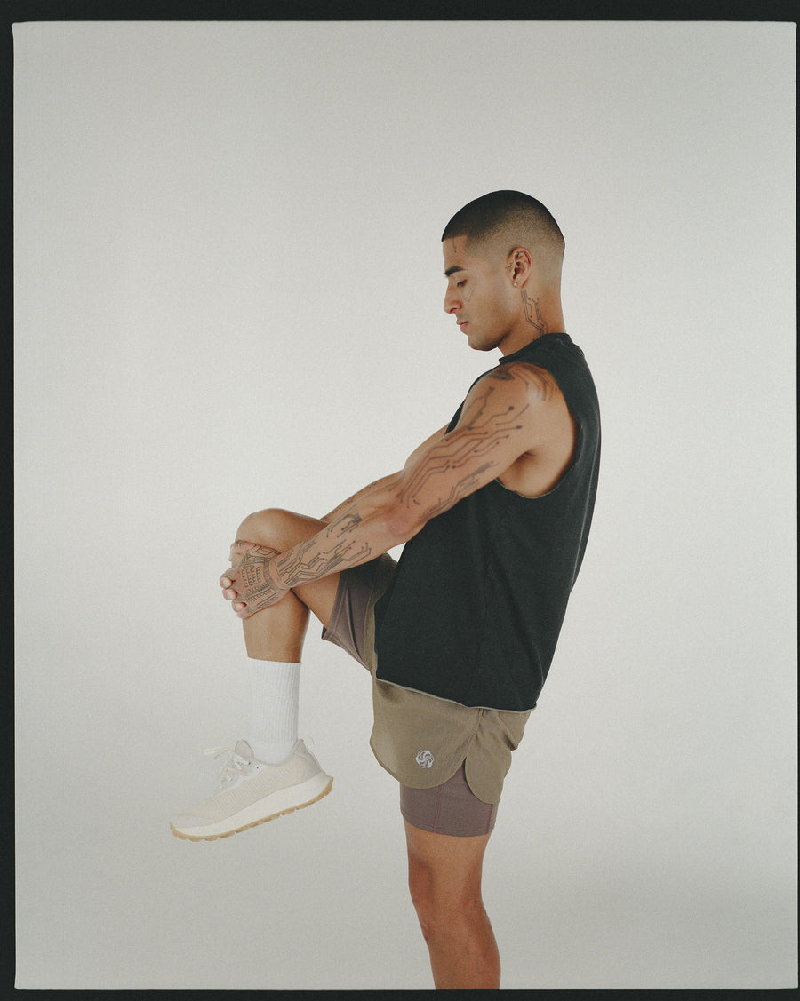 A man performing a knee hug stretch in a black tee and brown shorts.