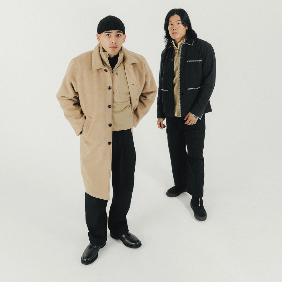 Two men - one in a tan long coat and the other in a black jacket - stand together looking at the camera.