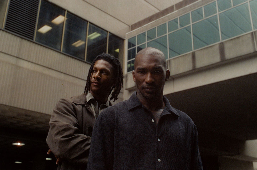 Two men pose in front of a concrete building.