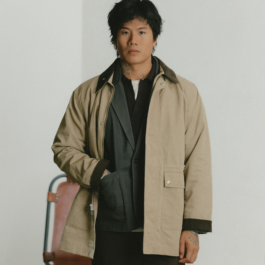 A man in a tan coat layered over a charcoal blazer with one hand in his pocket.