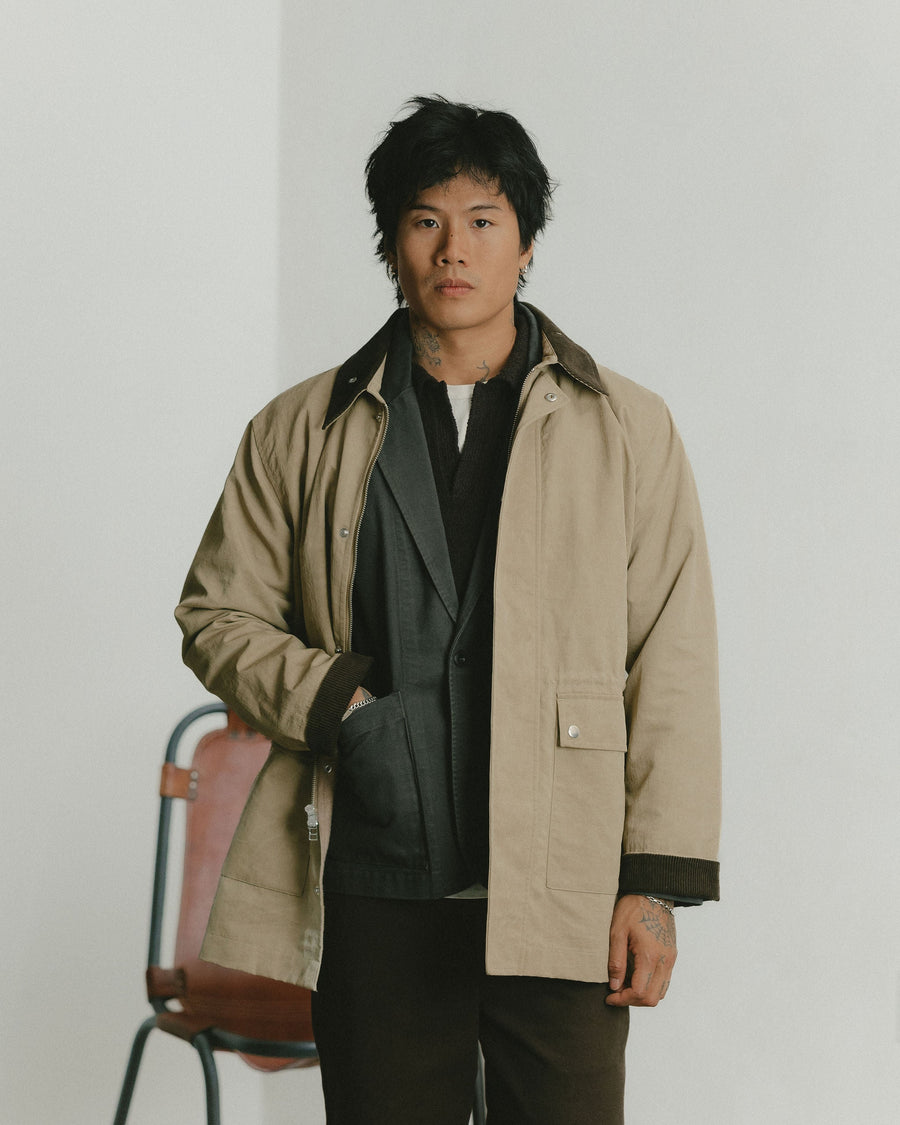 A man in a tan coat layered over a charcoal blazer with one hand in his pocket.