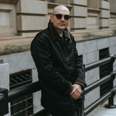 A man in black sunglasses and black nylon coat leans against some railing.