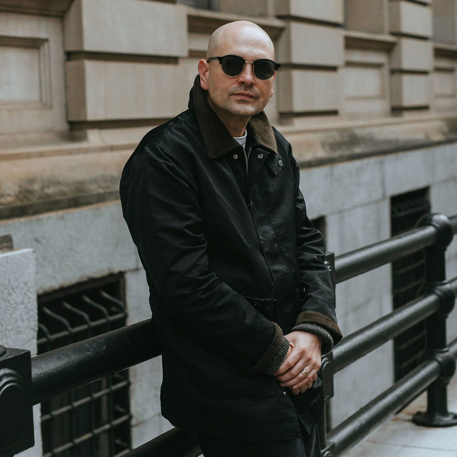 A man in black sunglasses and black nylon coat leans against some railing.