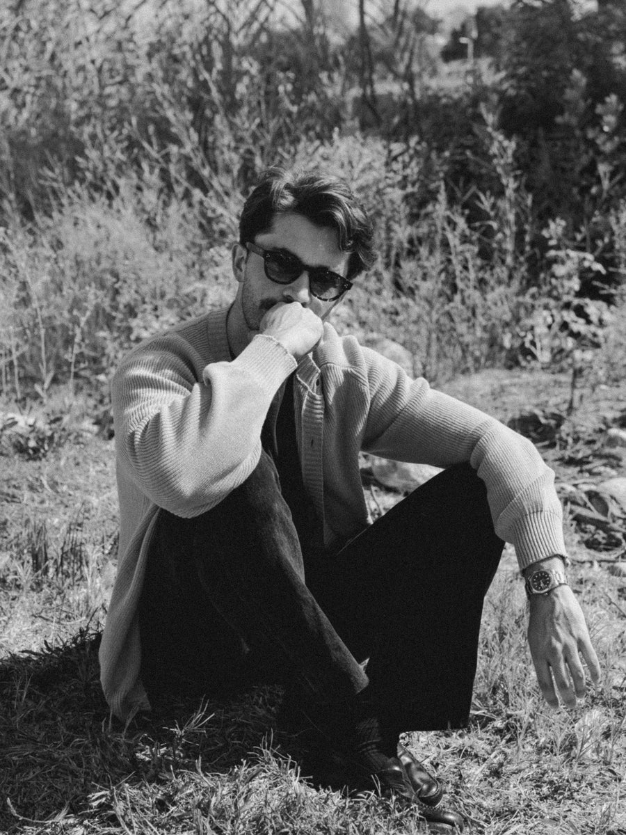 A man in sunglasses and a sweater sits on grass.