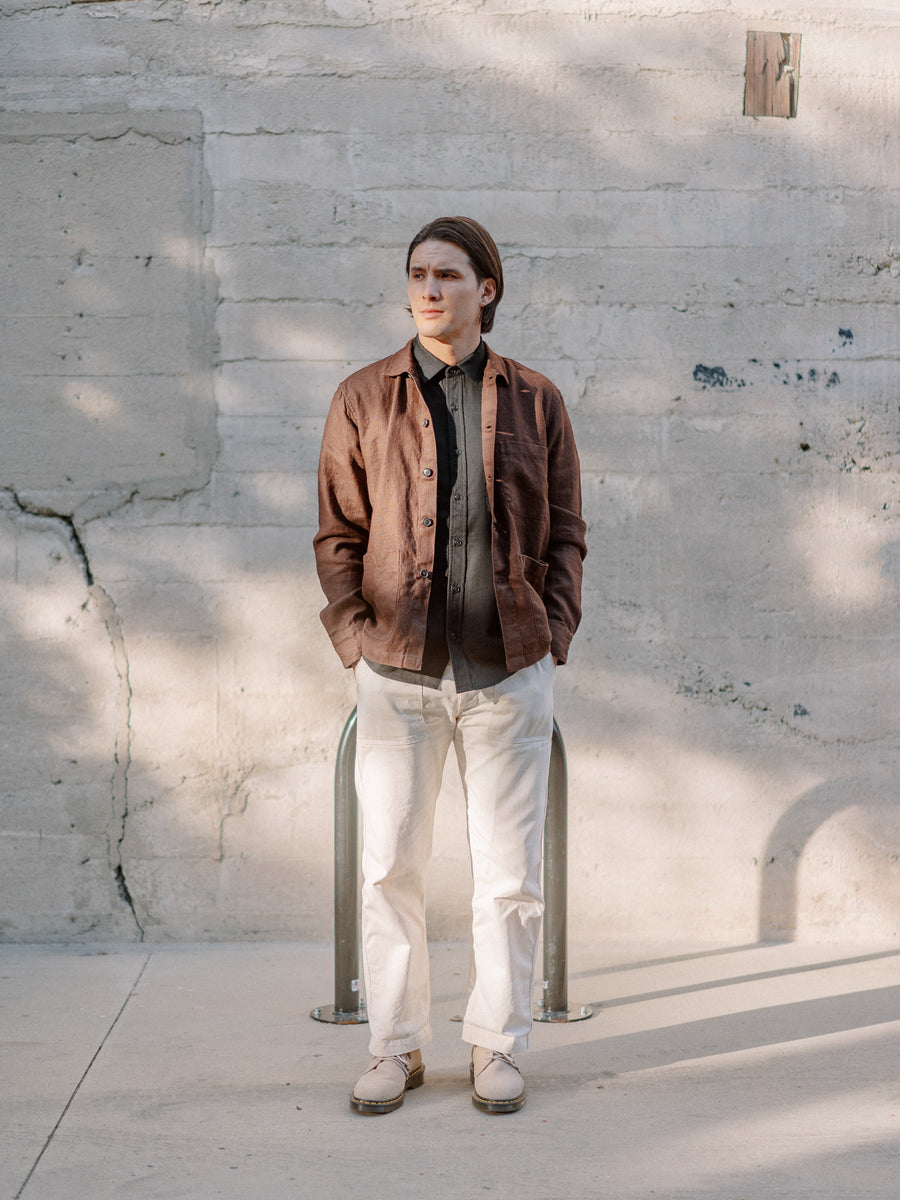 A man in a brown jacket and white pants stands in front of a concrete wall.
