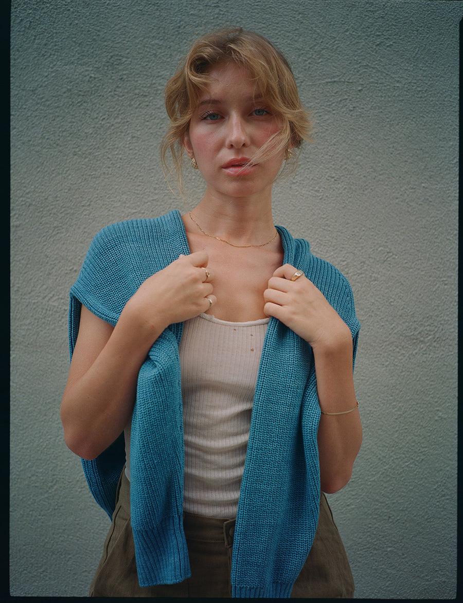A woman in a blue sweater stands in front of a grey wall