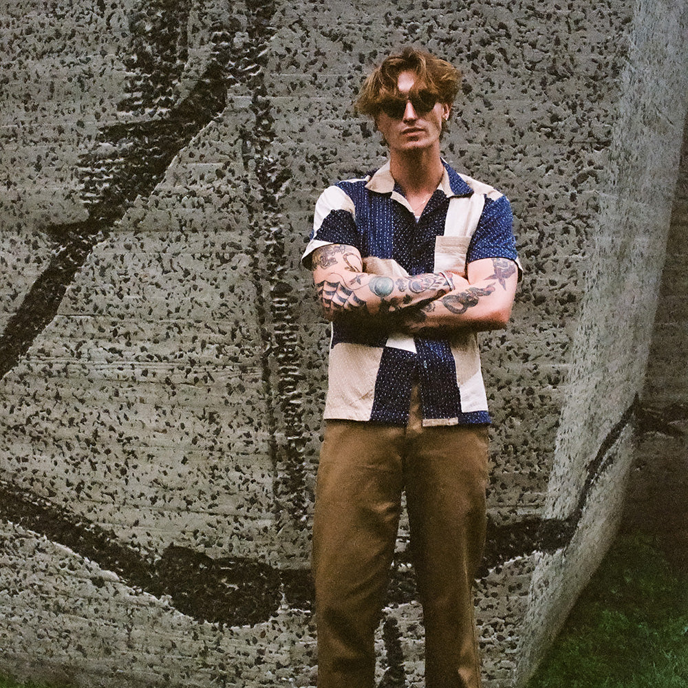 A man in sunglasses and a patchwork shirt stands against a concrete wall.