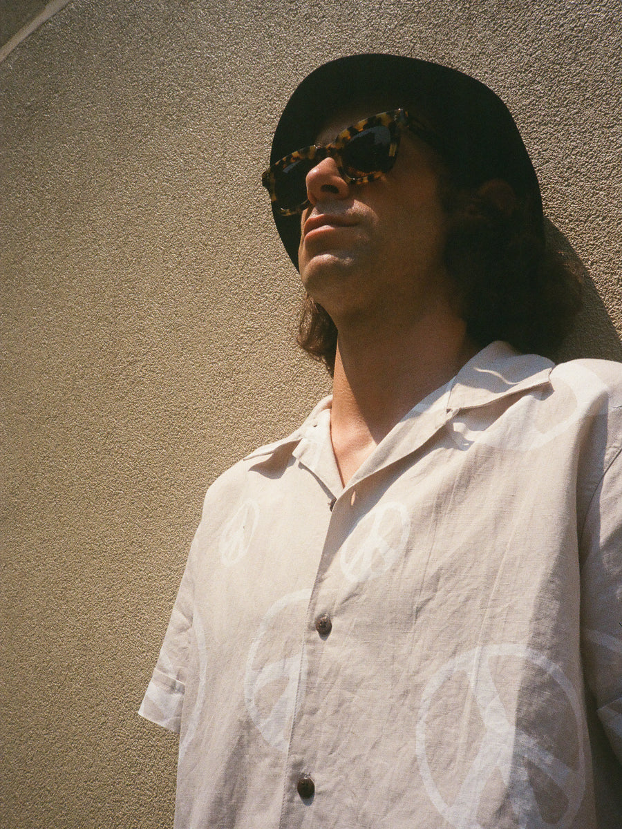 A man stands against a concrete wall wearing sunglasses, a black bucket hat and a shirt with white peace signs printed all over it.