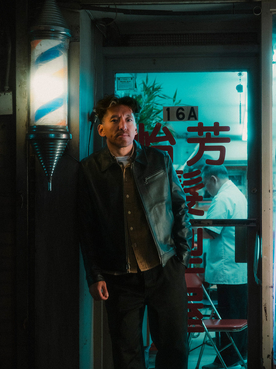 A man in a black jacket stands in front of a barbershop.