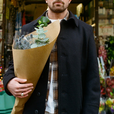 A man in a long black overcoat and brown flannel holds flowers in his arm