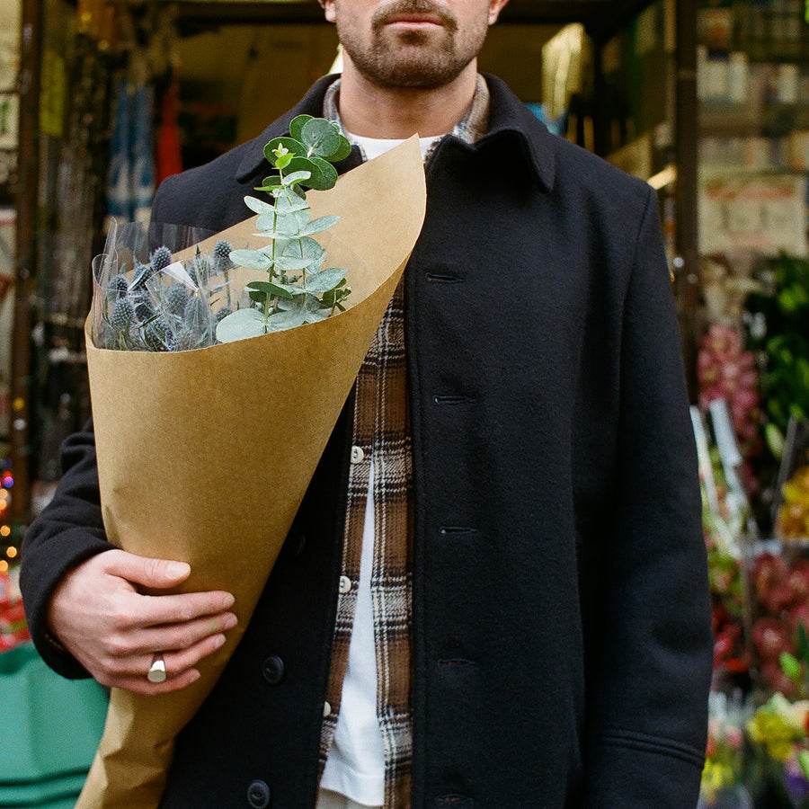 A man in a long black overcoat and brown flannel holds flowers in his arm