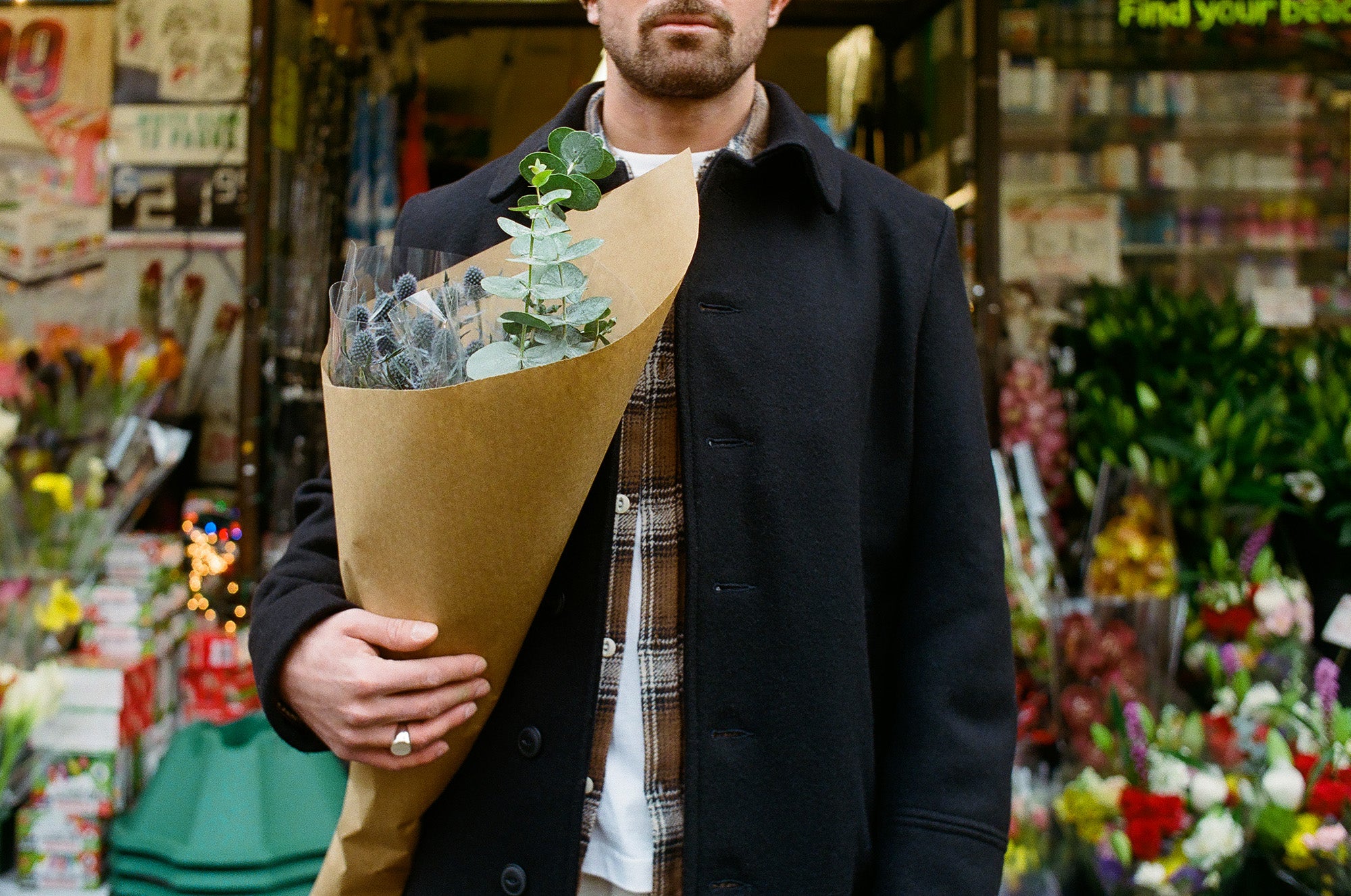 A man in a long black overcoat and brown flannel holds flowers in his arm