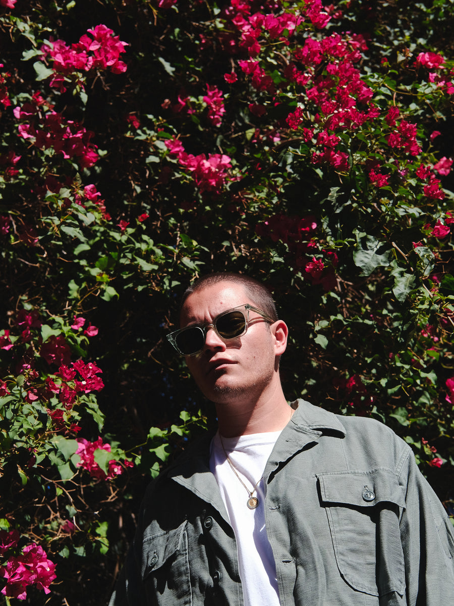 A man wearing sunglasses and an olive overshirt stands in front of pink flowers.