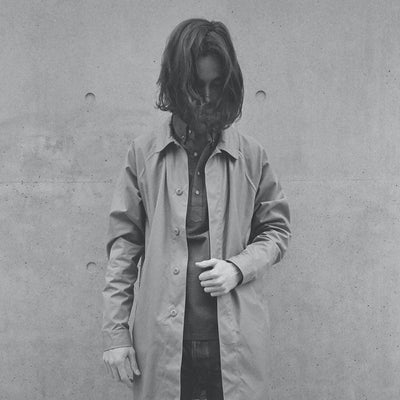 A man with his hair down wearing a light colored mac coat.
