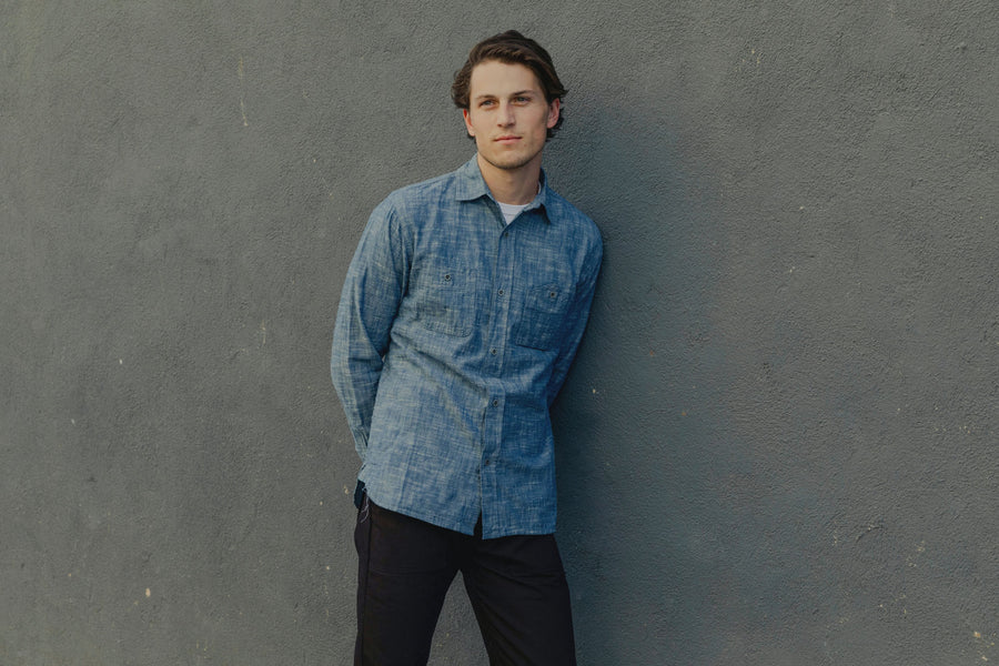 The model wears a light blue chambray 3sixteen shirt and leans against a wall, looking off camera.