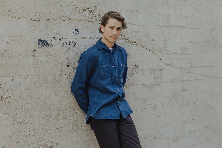 The model wears the deep indigo 3sixteen chambray and leans against a wall with his arms around his back.