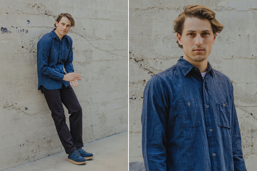The model wears a deep indigo chambray workshirt paired with black fatigue pants and blue suede shoes.