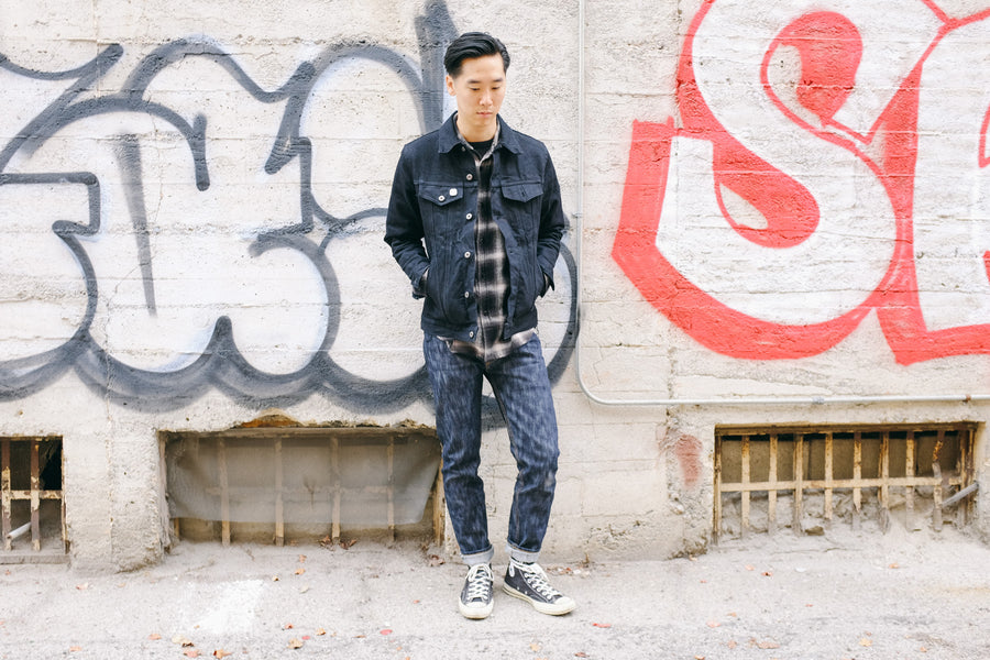 A model stands in front of a graffiti-adorned wall wearing a 3sixteen denim jacket, flannel and 15y anniversary denim.