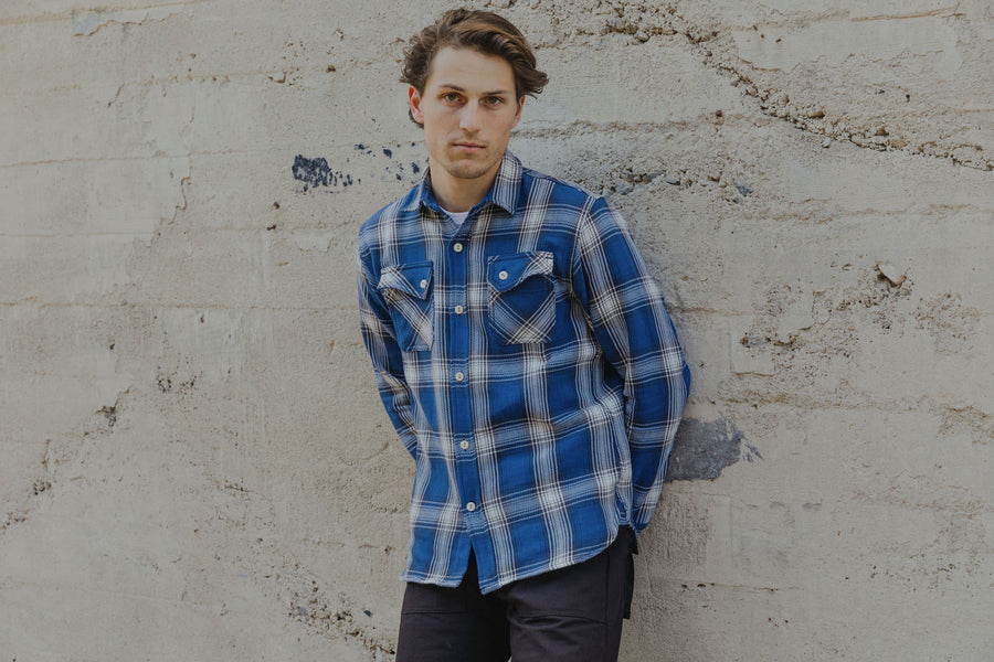 The model, dressed in an indigo check flannel by 3sixteen, leans against a concrete wall. 