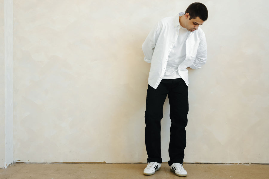 A man in a white shirt and black jeans stands in front of a cream, textured wall.