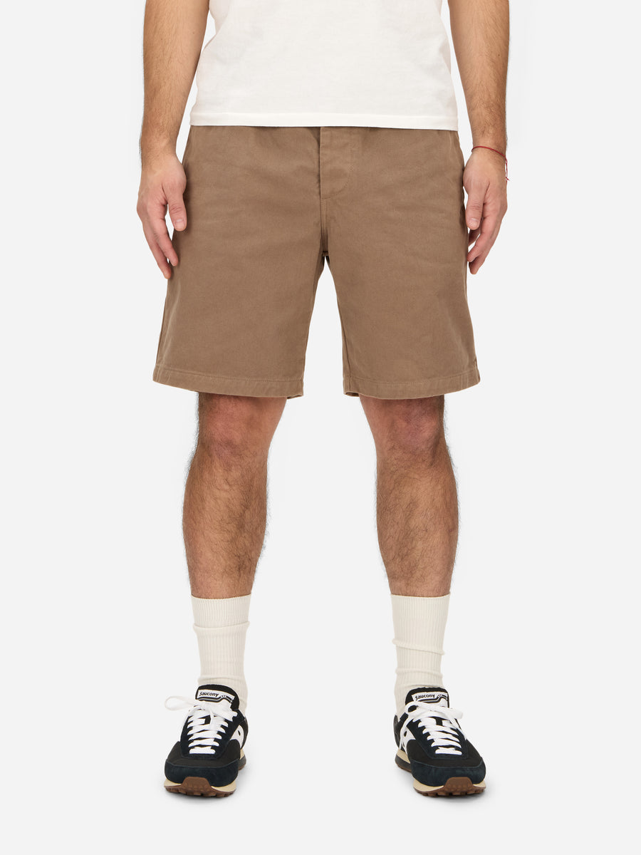 A person standing and wearing safari shorts in a beige color, with a relaxed fit and double pleated front, paired with a white t-shirt and low-top sneakers.