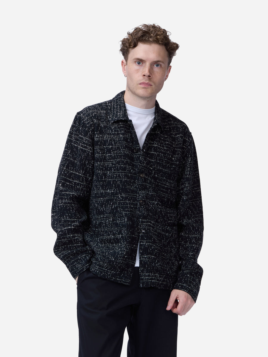 A man wearing a black and gray textured tweed jacket over a white t-shirt and black trousers, standing against a light gray background.