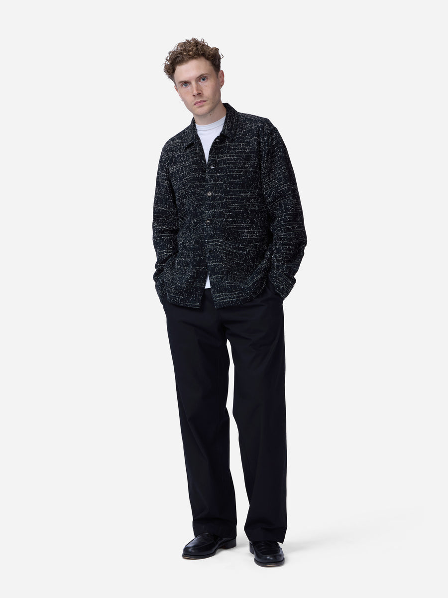 A man modeling a black and gray textured patterned jacket over a white t-shirt, paired with black trousers and black shoes, against a plain light gray background.