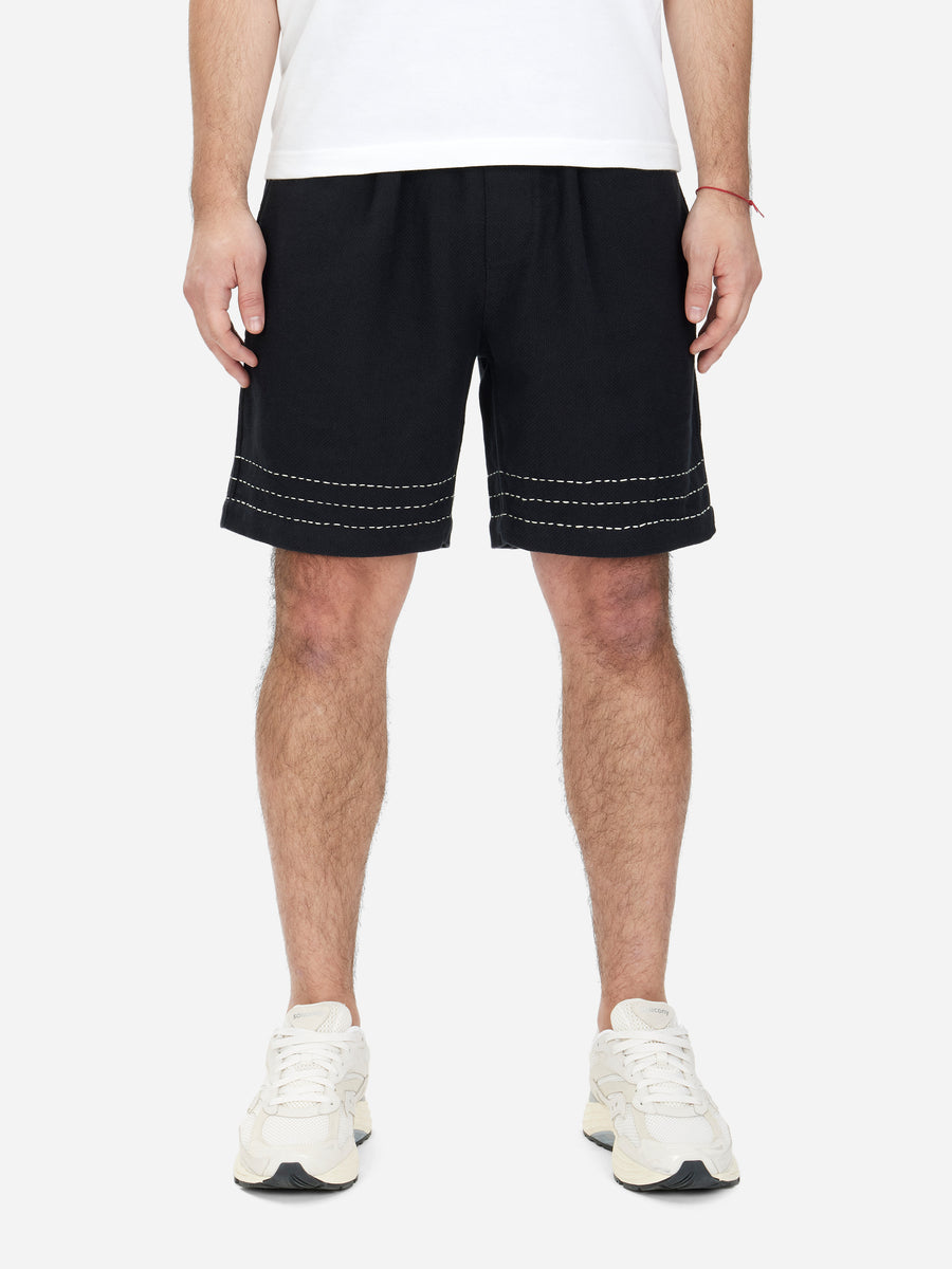 A pair of black soccer shorts with a relaxed fit, drawstring elastic waist, and hand stitching detail around the hem. Worn by a model with white sneakers.